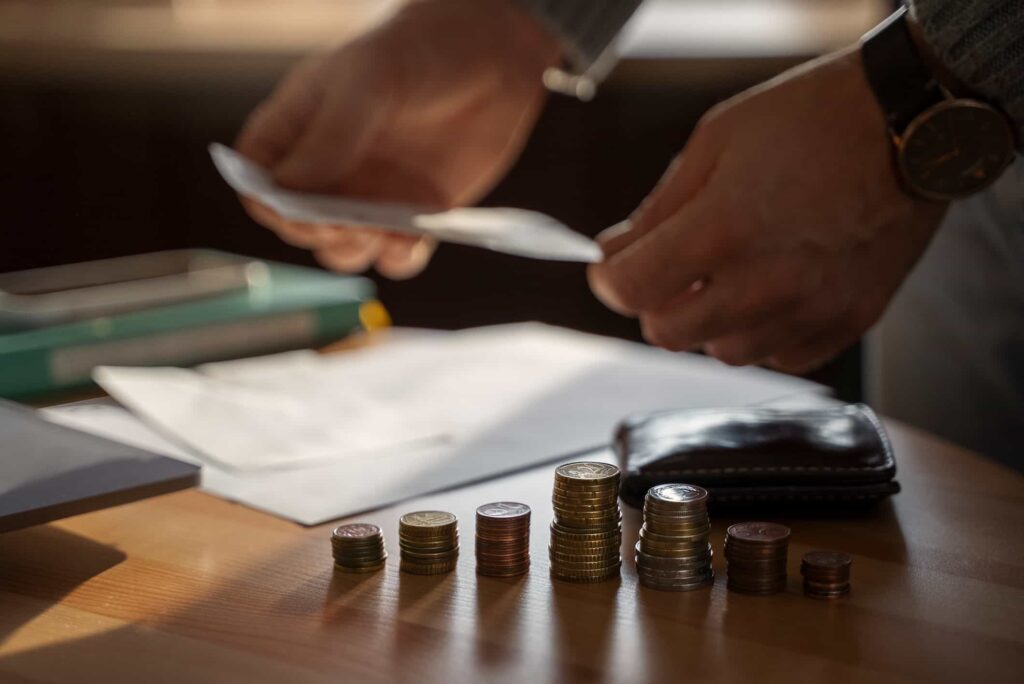 Stacked coins on the table with sheets of paper and books in the background, as well as two hands holding the paper.
