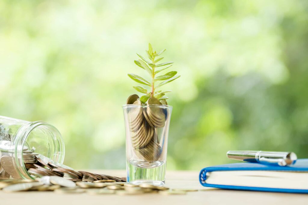 A glas filled with gold coins and a small plant, with a green background in nature.