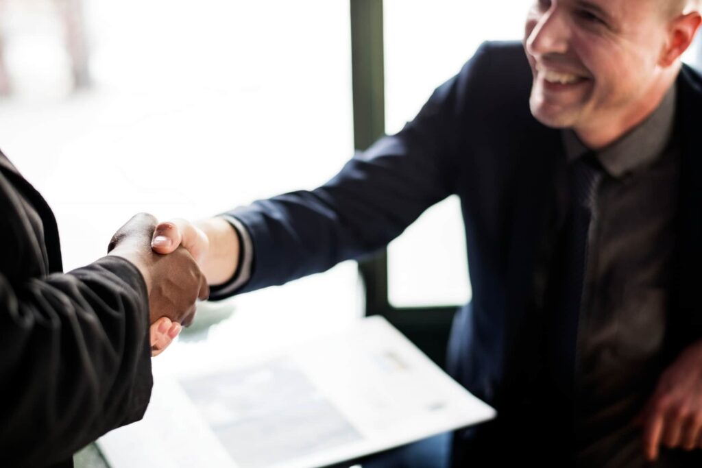 Two men in dark suits shaking hands, the man on the right side smiling happily.