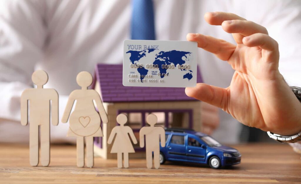 A man holding an international bank card beside a miniature wooden family, with a toy car and small model house in the background.