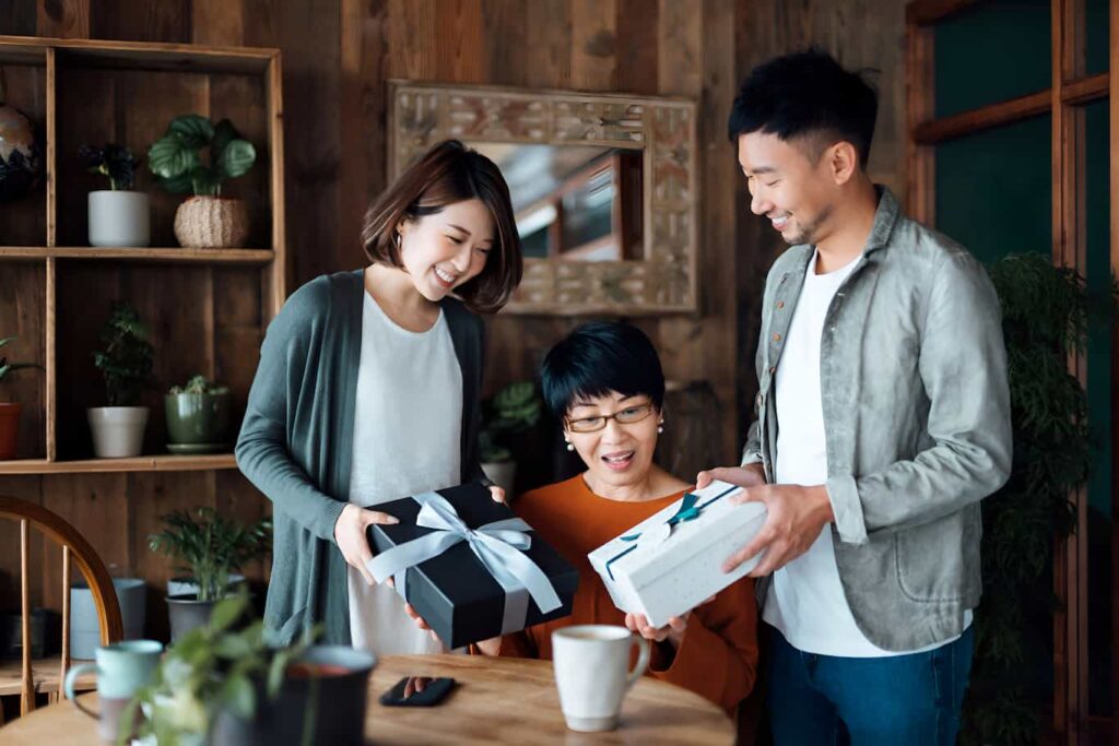 A couple holding out presents and smiling at a middle-aged woman seated between them, who looks surprised.